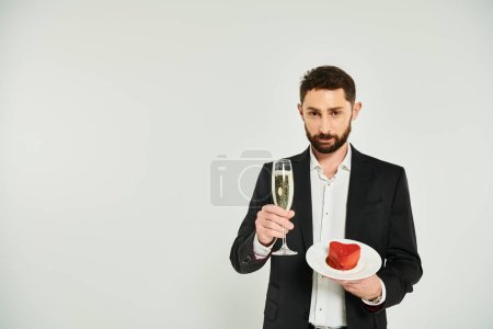 handsome elegant man with champagne glass and delicious heart-shaped cake looking at camera on grey