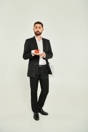 full length of sharp-dressed man holding plate with heart-shaped cake on grey, st valentines concept