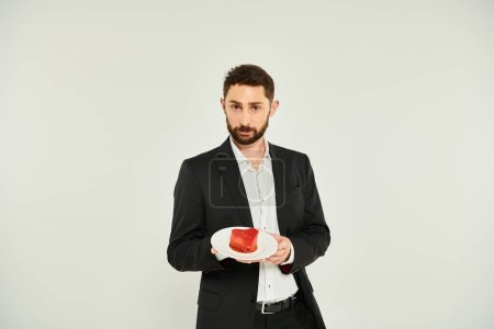 elegant bearded man showing plate with hear-shaped cake on grey, delicious Saint Valentines treat