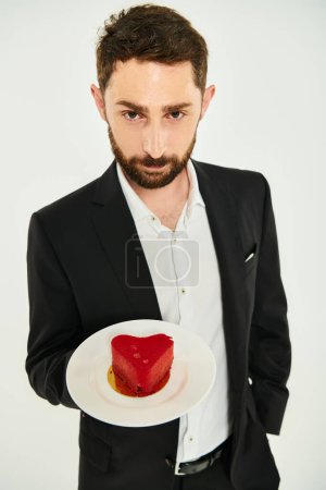 bärtiger eleganter Mann zeigt Teller mit rotem herzförmigen Kuchen auf grau, Valentinstag