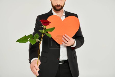 Ausgeschnittene Ansicht eines eleganten Mannes mit roter Rose und orangefarbenem Papierherz auf grau, Valentinskonzept