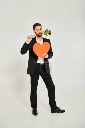 Valentinstag, gut aussehender Mann mit orangefarbenem Papierherz und roter Rose in Zähnen auf grau