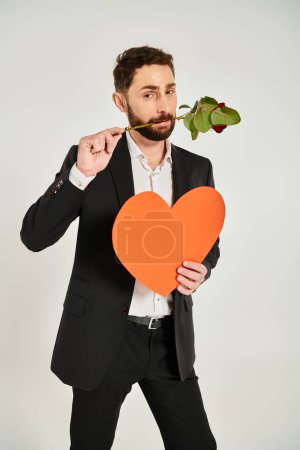 bärtiger stylischer Mann mit orangefarbenem Papierherz und roter Rose in den Zähnen auf grauem, st valentines Konzept