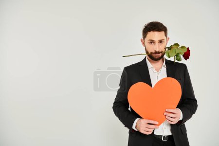 eleganter Mann mit riesigem Papierherz und roter Rose in den Zähnen, der am grauen Valentinstag in die Kamera blickt