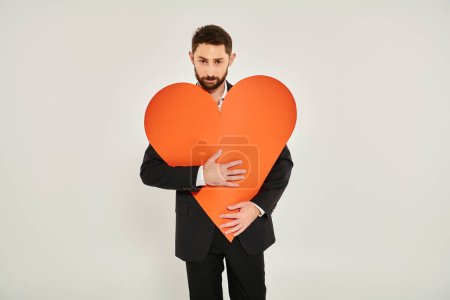 bearded charismatic man embracing huge paper heart looking at camera on grey, Saint Valentines day