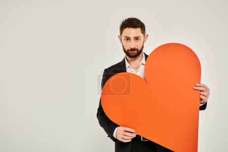 good-looking elegant man showing huge paper heart looking at camera on grey, St Valentines concept