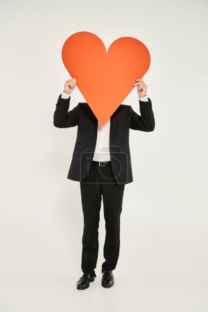 elegant man in black suit obscuring face with huge orange paper heart on grey, st valentines concept