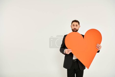well-dressed bearded man with huge orange paper heart looking at camera on grey, st valentines day