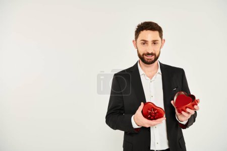 hombre barbudo sonriente en traje negro abriendo caja de regalo roja en forma de corazón en gris, regalo de San Valentín