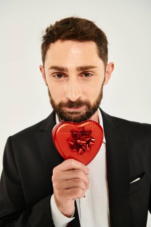 hombre barbudo feliz mostrando caja de regalo en forma de corazón con lazo rojo en gris, regalo de San Valentín