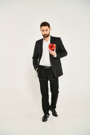 hombre seguro de sí mismo en traje negro sosteniendo la mano en el bolsillo y mostrando caja de regalo en forma de corazón rojo en gris
