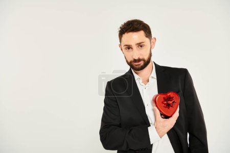 handsome elegant man with red heart-shaped Saint Valentines day present looking at camera on grey