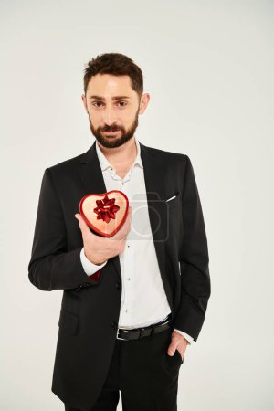hombre barbudo positivo y elegante mostrando caja de regalo en forma de corazón con arco en gris, día de San Valentín