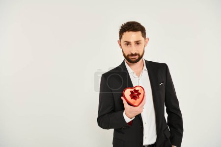 sonriente hombre carismático mostrando caja de regalo en forma de corazón con arco en gris, concepto de San Valentín