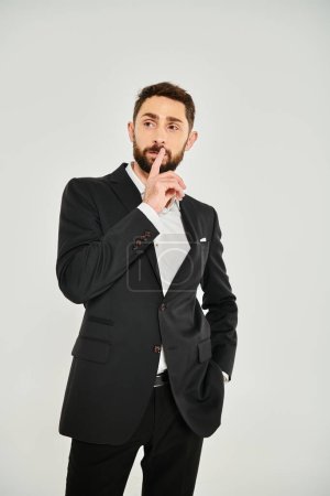 nachdenklicher Geschäftsmann im schwarzen Anzug, der Lippen berührt, während er mit der Hand in der Tasche auf grau steht