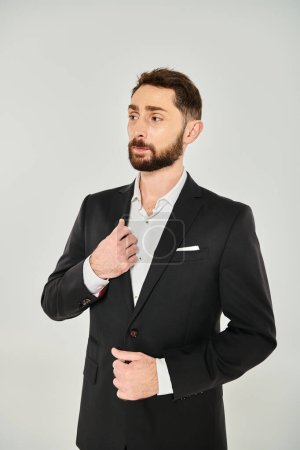nachdenklich eleganter bärtiger Geschäftsmann im schwarzen Anzug steht und vor grauem Hintergrund wegschaut