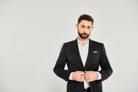 guapo barbudo hombre de negocios abotonando traje elegante negro y mirando a la cámara en el fondo gris