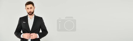 charismatic bearded businessman buttoning black suit and looking at camera on grey, banner