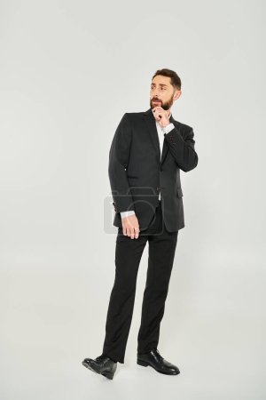 full length of serious elegant businessman in black suit touching beard and looking away on grey