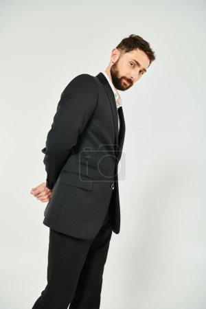 positive and elegant bearded businessman with hands behind back looking at camera on grey backdrop