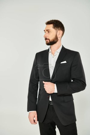 erfolgreicher bärtiger Geschäftsmann im schwarzen eleganten Anzug, der vor grauem Hintergrund wegschaut