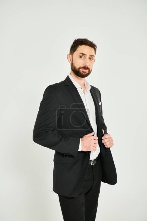 hombre de negocios carismático vestido agudo en ropa formal negro mirando a la cámara en el fondo gris
