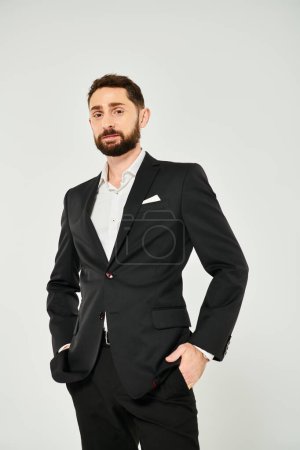 confident bearded businessman with hands in pockets looking at camera while posing on grey