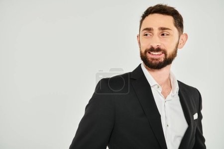 portrait of successful elegant bearded businessman in black suit smiling and looking away on grey