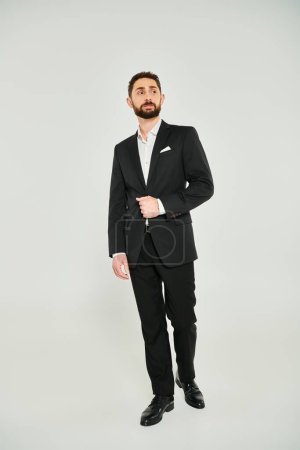 rico y exitoso hombre de negocios en traje negro de pie y mirando hacia otro lado en gris, longitud completa