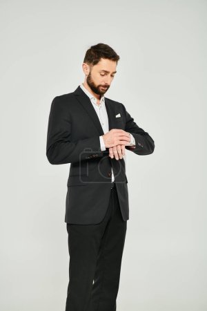 good-looking bearded businessman adjusting sleeve of black suit on grey backdrop in studio