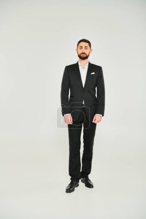 Vorderseite des bärtigen Geschäftsmannes im schwarzen eleganten Anzug, der vor grauem Hintergrund steht, in voller Länge