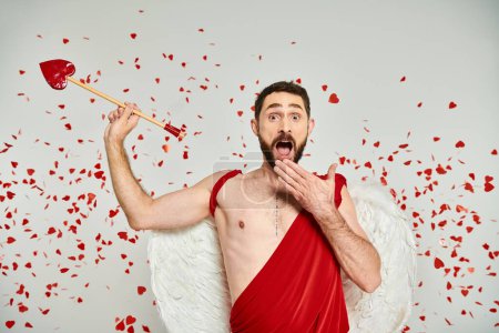 amazed bearded cupid man with arrow covering open mouth with hand under red heart-shaped confetti