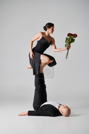 acrobatic woman in black attire holding red roses and balancing on feet of dancing partner on grey