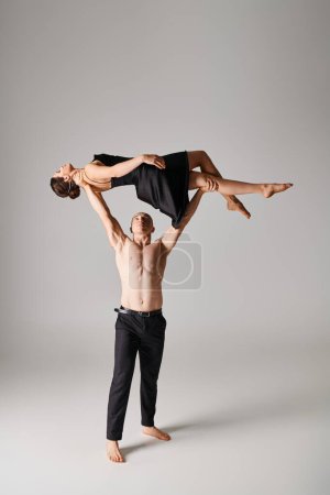 shirtless man lifting young woman in black dress while performing dress on grey background