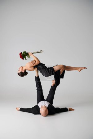 flexible woman in black attire holding red roses and balancing on feet of dancing partner on grey