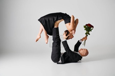 young flexible woman in black attire balancing on feet of dancing partner holding red roses on grey