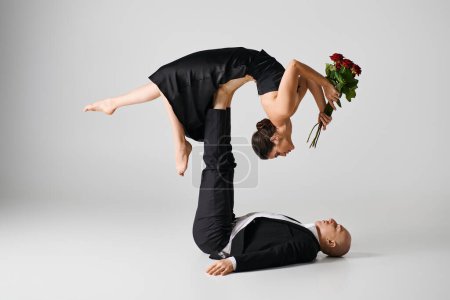 flexible Frau im schwarzen Kleid, rote Rosen in der Hand und balancierend auf den Füßen eines Tanzpartners auf grau