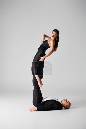 flexible young woman in black attire balancing on bare feet of athletic man on grey backdrop