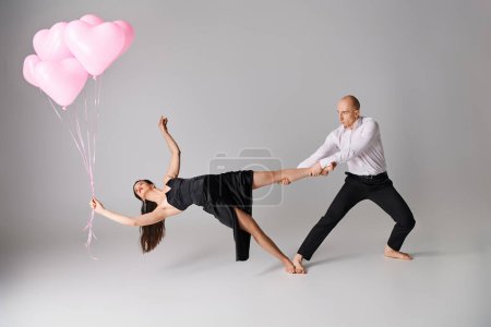 flexible junge Frau im schwarzen Kleid balanciert auf ihren nackten Füßen mit Luftballons in der Nähe von Mann auf Grau