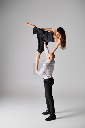 starker Mann hebt brünette Frau in schwarzem Kleid, während er vor grauem Hintergrund tanzt