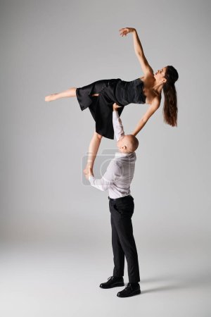strong man lifting flexible brunette woman in black dress while performing dance on grey backdrop