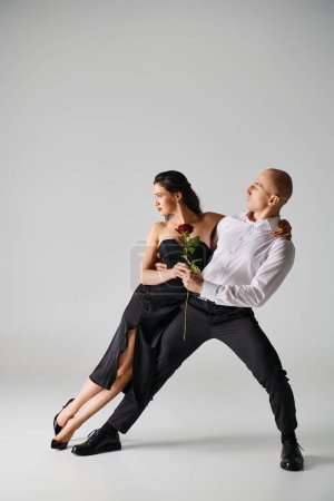 Elegant dance motion of young couple, woman holding red rose and man in formal attire in studio