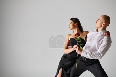 Dramatic dance motion of young couple, woman holding red rose and man in formal attire in studio