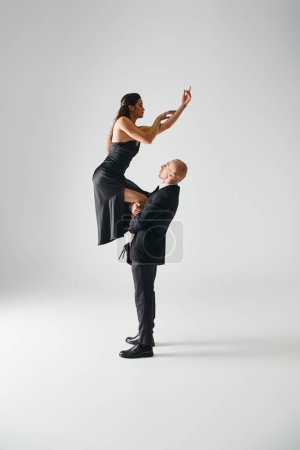 Foto de Hombre levantando joven morena bailarina femenina en vestido negro y tacones altos equilibrio durante el rendimiento - Imagen libre de derechos