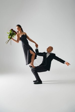 hombre levantando mujer joven elegante en vestido negro sosteniendo rosas rojas y equilibrio durante el rendimiento