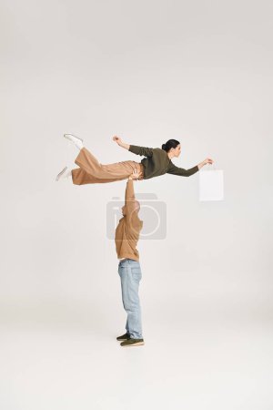 strong man in casual attire lifting woman with shopping bag, balancing in a playful pose in studio
