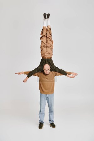 strong man in casual wear supporting body of acrobat partner while rehearsing in studio on grey