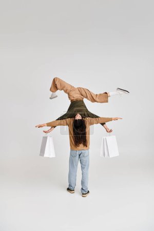 acrobatic woman holding purchases and balancing upside down with support of partner on grey