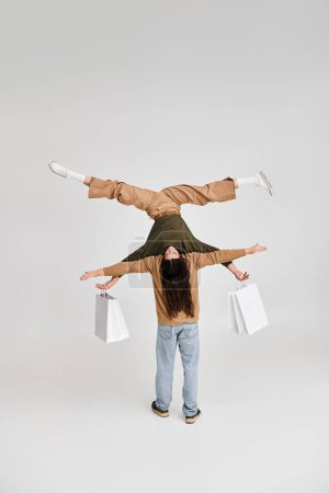 Frau hält Einkaufstüten und balanciert kopfüber mit Unterstützung ihres akrobatischen Partners auf grau