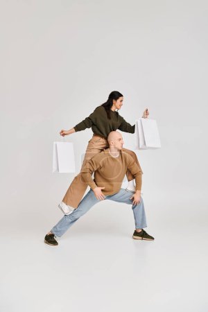 acrobatic performance of young couple, woman with shopping bags balancing on body of man on grey